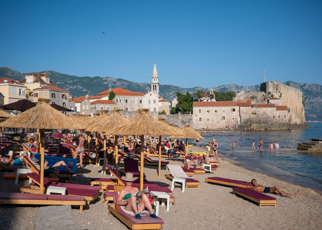 Crowded Plaza Ricardova Glava beach with lounge chairs and Budva Old Town in the background.