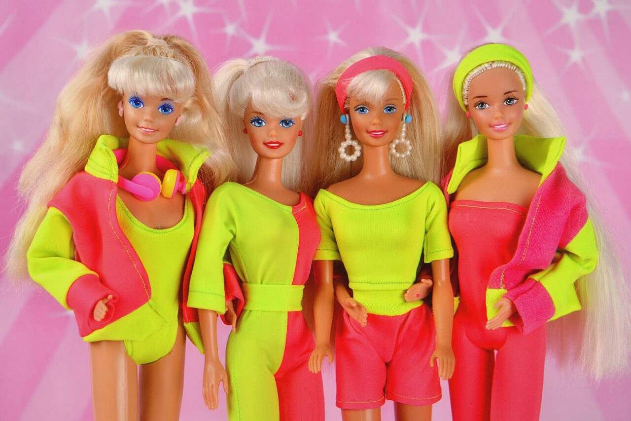 Four barbie dolls in neon clothing on a pink background