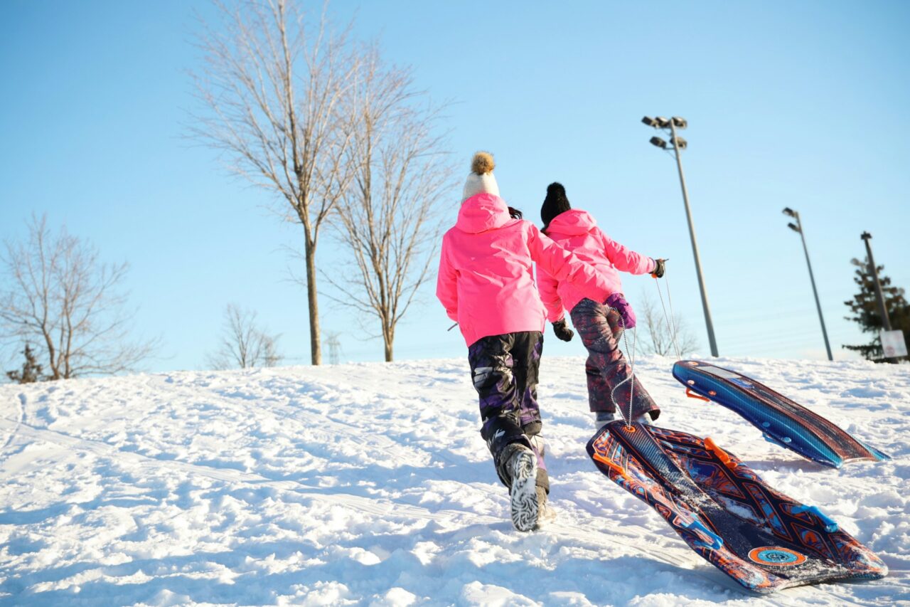 Two kids with pink jackets and toboggans walking up a snowy hill