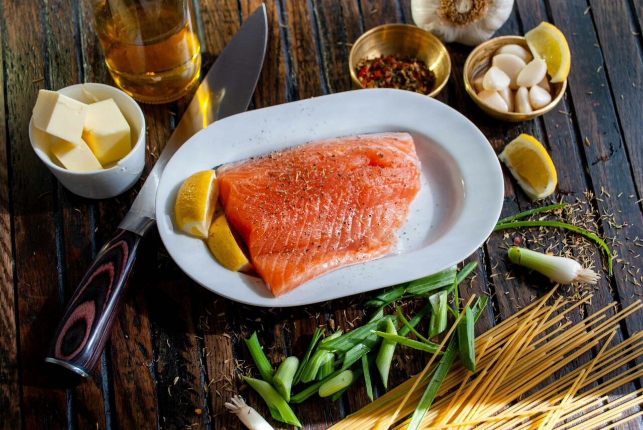 Raw fillet of salmon on a white plate surrounded by a knife, butter, garlic and spring onions