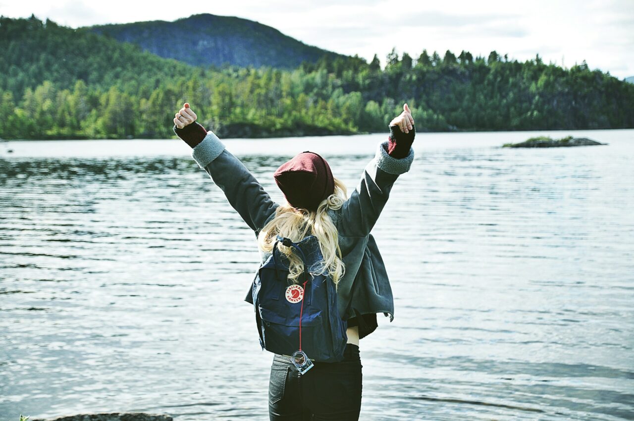 Woman with a backpack standing in front of a lake raising arms in the air