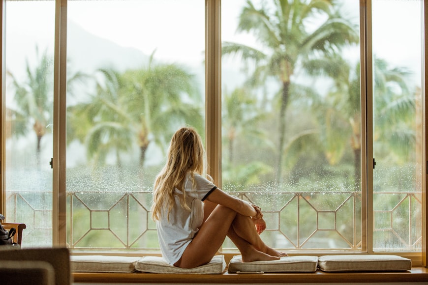 Woman with long blonde hair looking out of a window at palm trees
