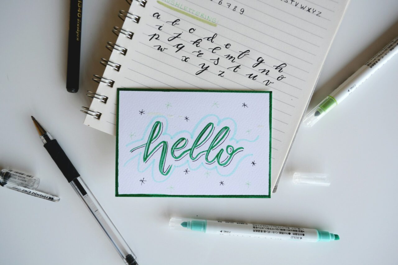 Hello written on a card on top of a notebook surrounded by pens